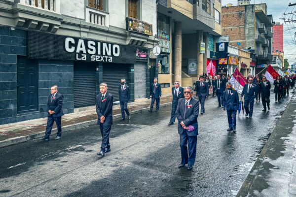 Dignitaries march in formal attire along a wet street in Ipiales, Nariño, Colombia, carrying flags during a solemn procession.