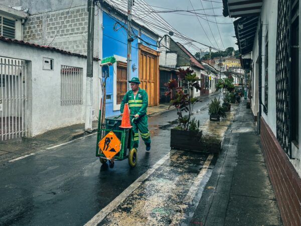 A street worker in a green uniform pushes a cart along a rainy, colorful neighborhood in Cali, Valle del Cauca, Colombia.