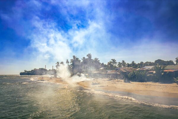Gentle waves lap at the sandy shore of Juanchaco, Valle del Cauca, with palm trees and mist rising against a vibrant blue sky.