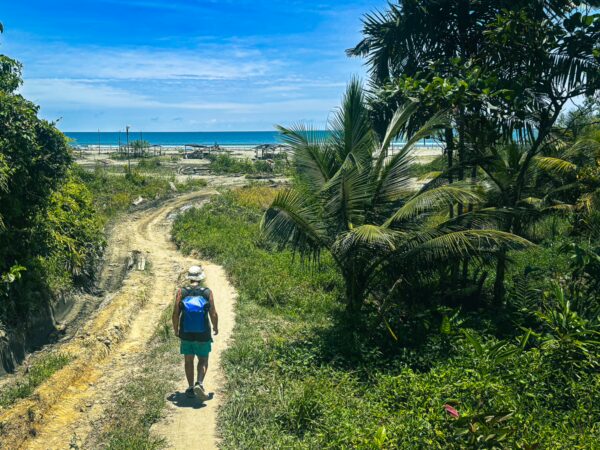 A traveler walks along a sandy path lined with lush greenery, leading to the blue waters of La Barra in Valle del Cauca, Colombia.