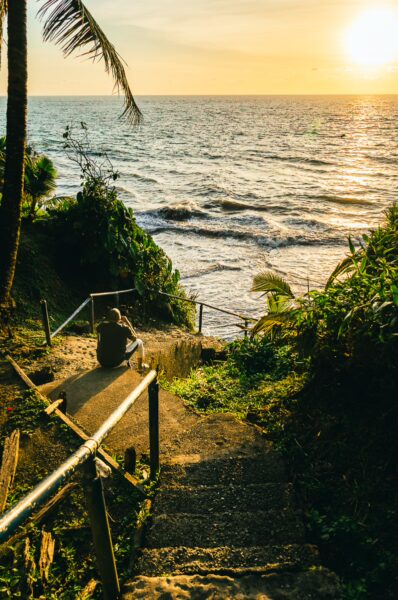 A person sits on steps leading to the ocean at Ladrilleros, Valle del Cauca, Colombia, with a vibrant sunset over the waves.
