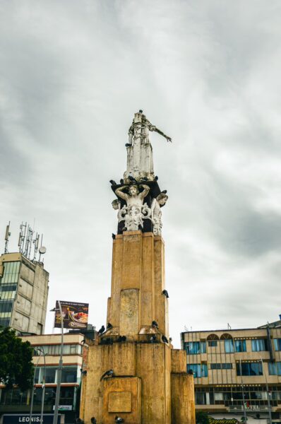 Monument with a large statue atop a tall pedestal surrounded by urban buildings under a cloudy sky in La Pola, Ipiales, Nariño, Colombia.