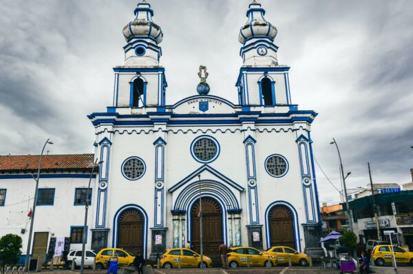 Iglesia San Felipe Neri features striking blue and white architecture with twin towers in Ipiales, Nariño, Colombia. Yellow taxis park nearby.