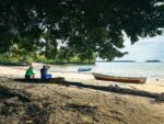 Relaxing at Juan de Dios Beach in Uramba, a tranquil scene with two people sitting under trees by the calm waters of Valle del Cauca.