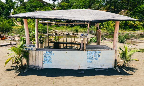 Colorful beach hut with a thatched roof on sandy shores of La Barra, Valle del Cauca, surrounded by lush greenery.