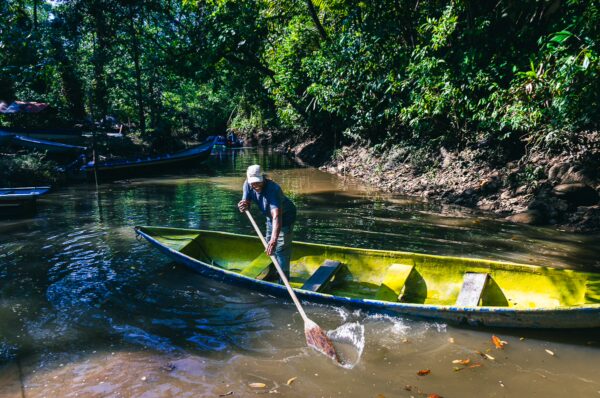 A person paddles a vibrant yellow canoe through the calm waters of Piscina Natural Pachenda, surrounded by lush green foliage in Ladrilleros, Valle del Cauca, Colombia.