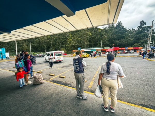 Travelers and locals gather at Rumichaca border in Nariño, Colombia, with shops and vehicles in the background under a cloudy sky.