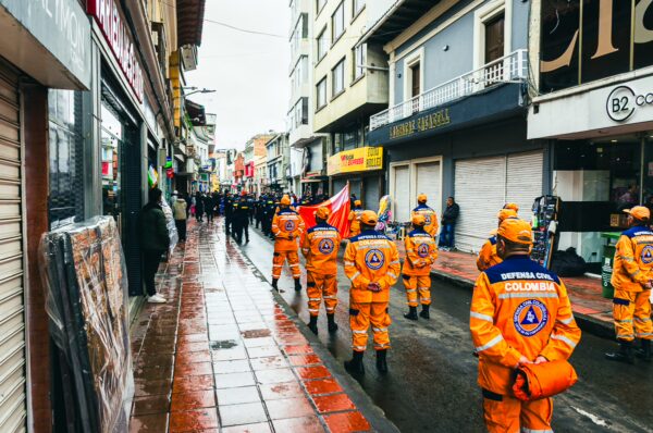 Emergency responders in orange uniforms march along a wet street in Ipiales, Narino, Colombia, surrounded by colorful storefronts.