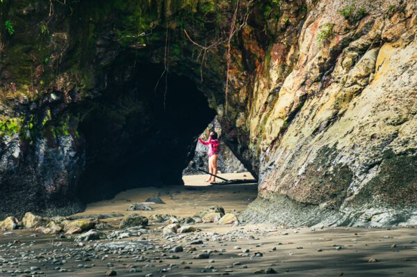 Waves crash against rocky cliffs at Juan de Dios Beach, with a person standing under a natural stone arch framed by lush greenery.