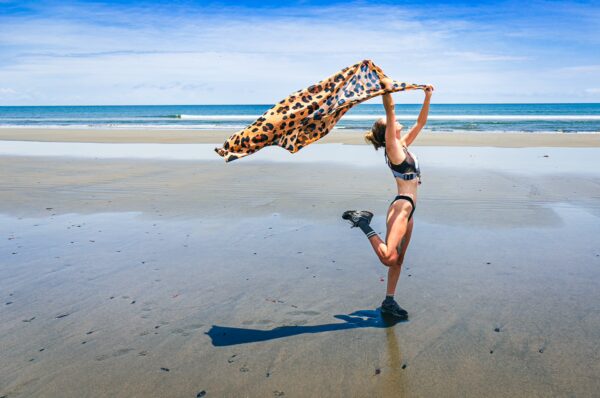 A woman in a bikini joyfully embraces a colorful scarf while dancing on the sandy beach of La Barra, Valle del Cauca, Colombia.