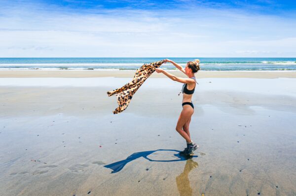 A woman in a bikini playfully waves a patterned scarf on the sandy beach of La Barra, Valle del Cauca, Colombia, under a blue sky.