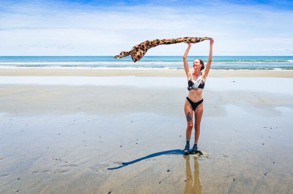 A woman in a bikini stands on the wet sand of La Barra, Valle del Cauca, Colombia, playfully holding a patterned scarf against the ocean backdrop.