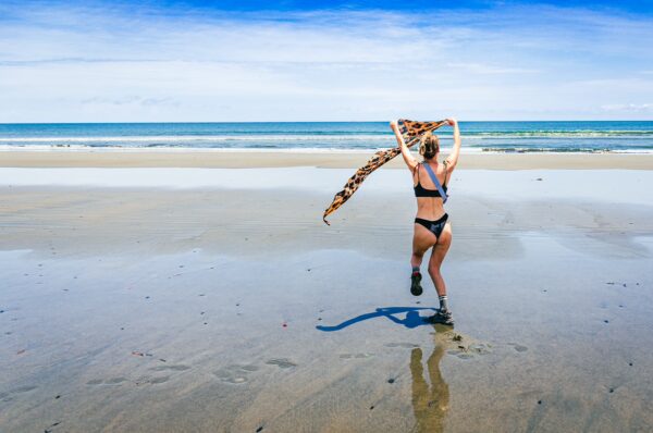 A woman in a bikini runs along the sandy beach, holding a patterned scarf against the vibrant blue sky at La Barra, Valle del Cauca, Colombia.