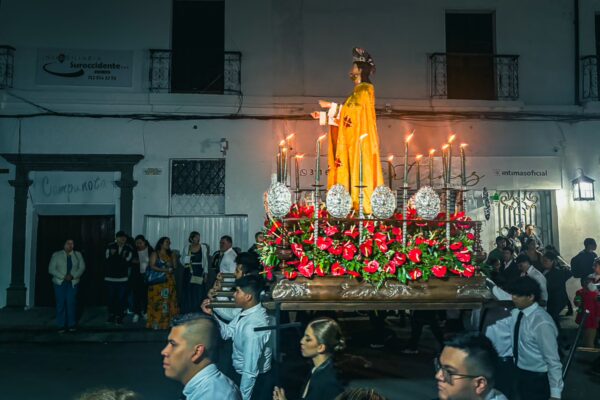 A vibrant procession in Popayán, Colombia, featuring a statue adorned with flowers and candles, surrounded by onlookers in traditional attire.