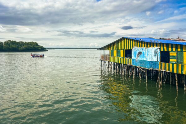 Brightly painted wooden house on stilts beside calm waters in Buenaventura, Valle del Cauca, Colombia, with a boat in the distance.