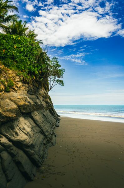 Lush greenery atop rocky cliffs meets the serene sandy shore at Juan de Dios Beach, Uramba, Valle del Cauca, Colombia.