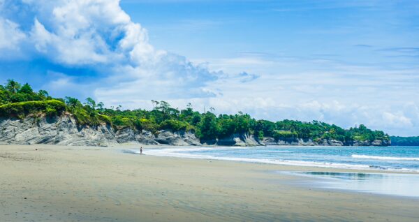 Soft sandy beach meets gentle waves, framed by lush green cliffs under a bright sky in La Barra, Valle del Cauca, Colombia.