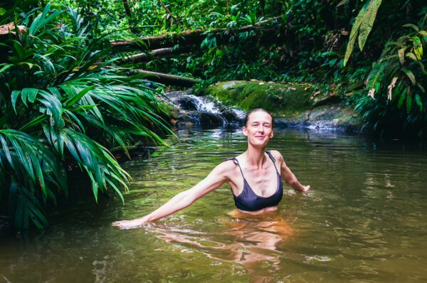 A woman smiles while enjoying a swim in a lush green pool surrounded by vibrant foliage and gentle waterfalls in Tres Marias, Uramba, Colombia.