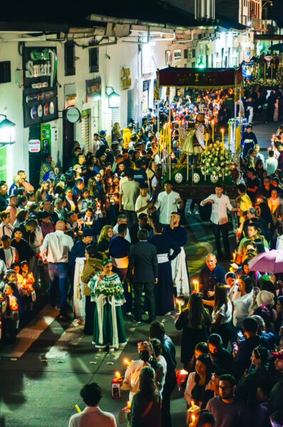 Colorful procession in Popayán, Cauca, Colombia, with crowds holding candles and traditional attire, illuminated by street lights.