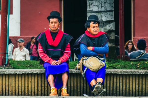 Two men in traditional clothing sit on a bench at Parque Principal Silva, surrounded by vibrant colors and local visitors in Cauca, Colombia.