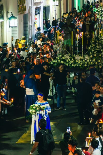 Vibrant street scene in Popayán, Cauca, Colombia, showcasing a procession with participants holding candles and flowers amidst a large crowd.