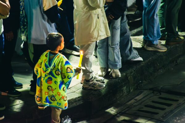 A child in a colorful poncho holds a candle while standing among a crowd during a nighttime event in Popayán, Cauca, Colombia.