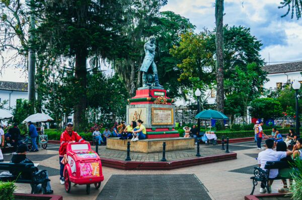 Colorful statue in Parque Caldas, Popayán, with people gathered around, surrounded by trees and benches under a cloudy sky.