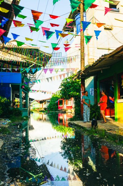 Colorful flags decorate a vibrant street in Juanchaco, Valle del Cauca, reflecting in a nearby puddle under blue skies.