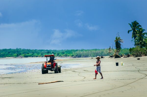 Tractor on the sandy beach at La Barra, Valle del Cauca, Colombia, with a person walking along the shoreline under blue skies.