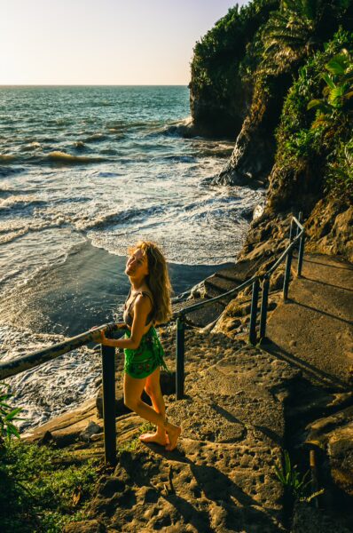 A girl in a green outfit stands by the ocean, overlooking rocky cliffs and gentle waves at Ladrilleros, Valle del Cauca, Colombia.