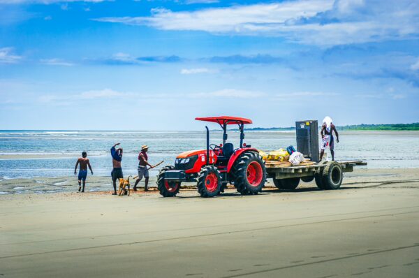 A vibrant beach in La Barra, Valle del Cauca, Colombia, features a red tractor on the sand with locals working nearby.