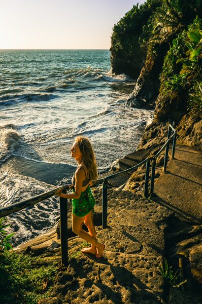 Golden sunlight illuminates a girl in green swimwear gazing at the waves along the rocky shore of Ladrilleros, Valle del Cauca, Colombia.