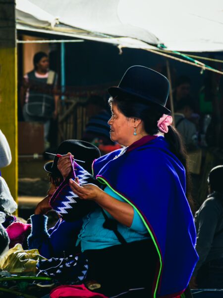 Brightly dressed woman in a blue poncho engages with textiles at the bustling market in Silva, Cauca, Colombia.