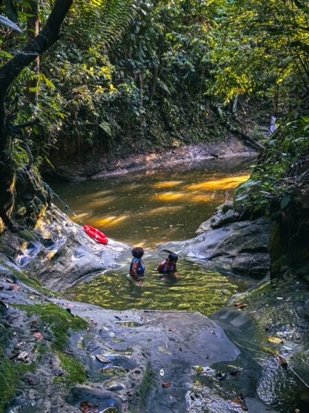 Two people enjoy a natural pool surrounded by lush greenery and rocky terrain at Piscina Natural Pachenda, Ladrilleros, Valle del Cauca, Colombia.