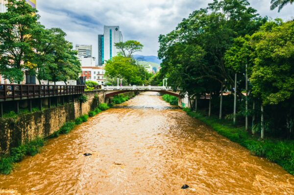 Flowing brown river lined with lush greenery and urban buildings in Cali, Valle del Cauca, Colombia. Overcast sky enhances the scene.