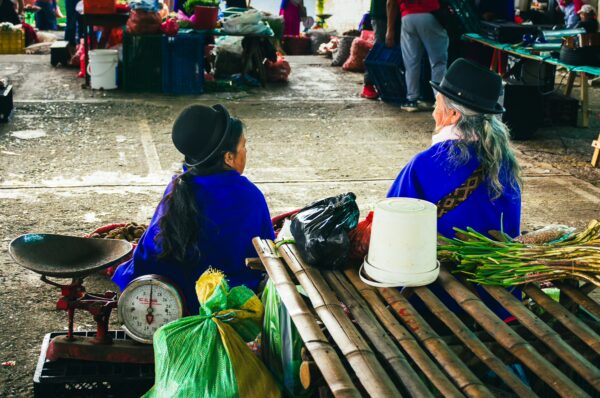 Local vendors wearing traditional blue attire sit by a scale amidst colorful market goods in Cauca, Colombia.
