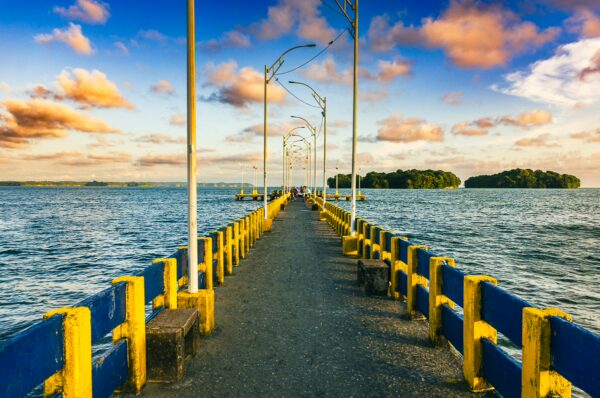 Vibrant blue and yellow pier stretches over calm waters in Juanchaco, Valle del Cauca, Colombia, under a colorful sunset sky.