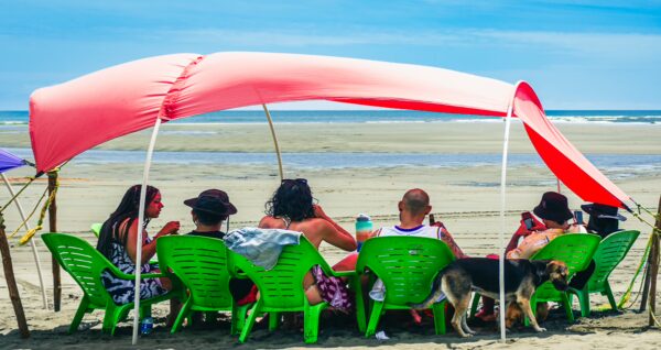 Bright green chairs under a red canopy on a sandy beach, with a group of people relaxing by the ocean in Valle del Cauca, Colombia.