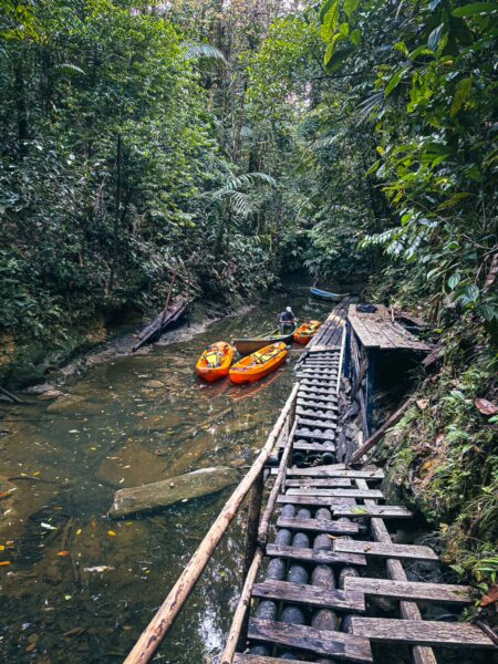 Bright orange kayaks rest along a wooden path beside a serene, lush river in the natural pool area of Ladrilleros, Valle del Cauca, Colombia.