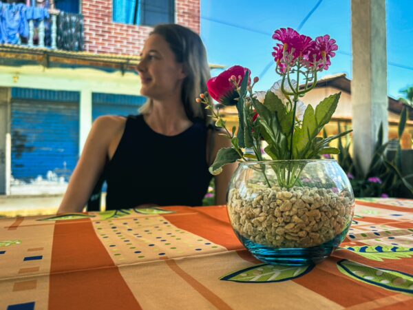 Vibrant flowers in a glass vase on a table, with a woman seated nearby in a colorful street of Ladrilleros, Valle del Cauca, Colombia.