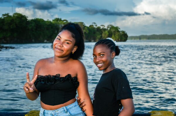 Two girls smile and pose by the water in Juanchaco, with lush greenery and a serene river setting in Valle del Cauca, Colombia.