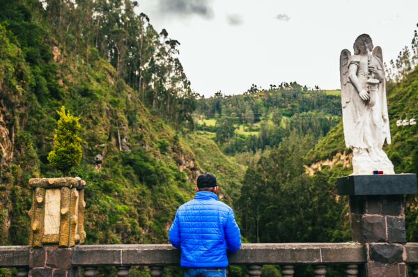 A traveler in a blue jacket stands by a stone railing, overlooking the lush valley of Las Lajas, Colombia. A white angel statue is nearby.