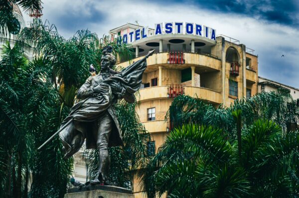 Statue with an umbrella stands in front of the Astoria Hotel, surrounded by lush palm trees in Plaza de Cayzedo, Cali, Valle del Cauca.