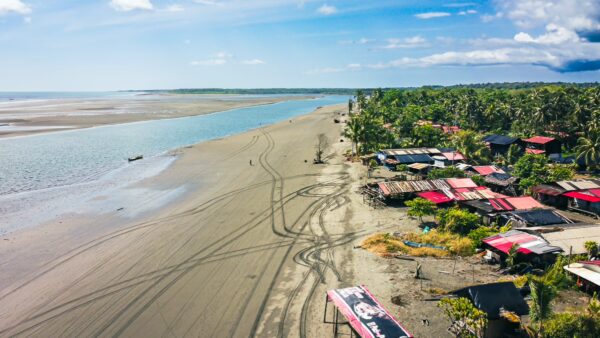 Vast sandy beach lined with colorful huts and palm trees, with a river flowing in the distance at La Barra, Valle del Cauca, Colombia.