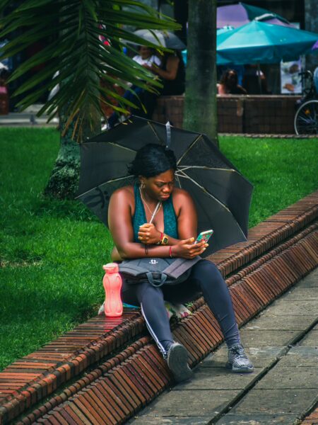 Relaxing in Plaza de Cayzedo, a woman sits under an umbrella, enjoying her phone amidst lush greenery and vibrant colors in Cali, Valle del Cauca.