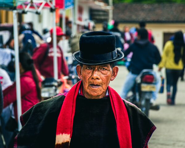 A man in traditional attire with a black hat and red scarf stands amidst a bustling street scene in Cauca, Colombia.