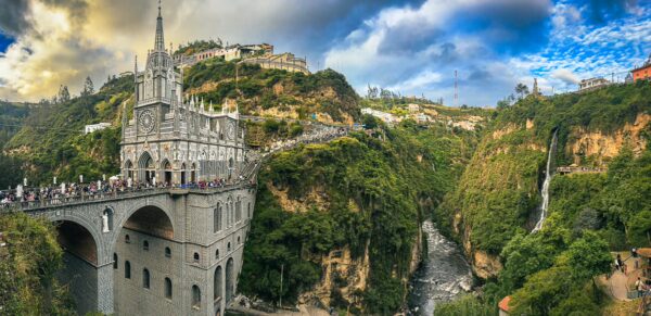 Lush green hills frame the stunning Las Lajas Sanctuary, nestled in a dramatic canyon in Narino, Colombia, with a cascading waterfall nearby.