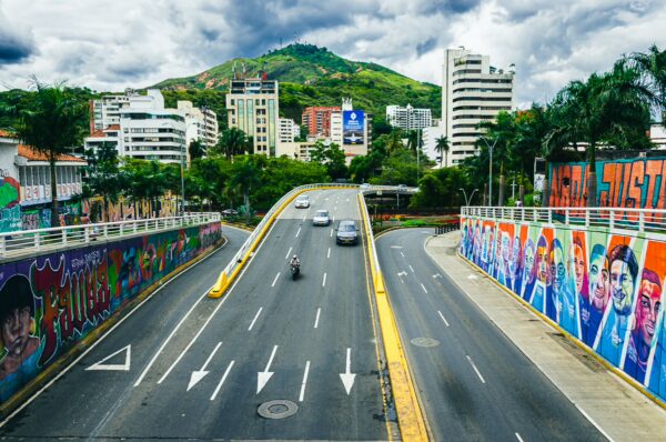 Vibrant street art decorates the walls along the road in San Antonio, Cali, with green hills and modern buildings in the background.