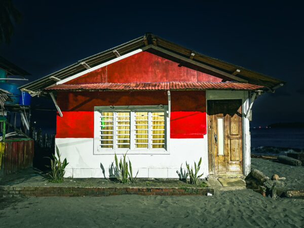 Bright red and white house with wooden door stands on the beach in Juanchaco, Valle del Cauca, Colombia, under a night sky.