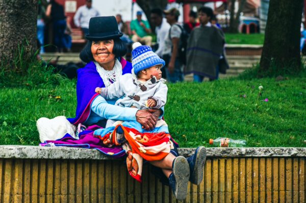 Traditional woman in vibrant clothing holds a child, seated on a bench in Parque Principal Silva, Cauca, Colombia, surrounded by greenery.